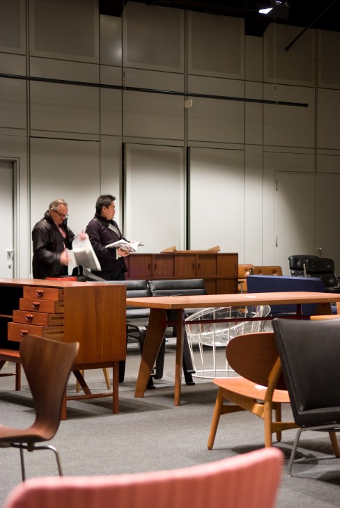 Two men inspect vintage furniture at an auction, surrounded by mid-century chairs, tables and dressers.