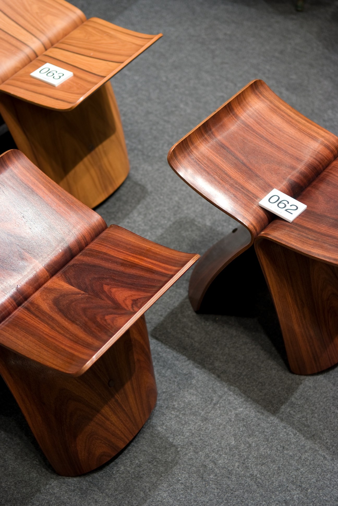 Wooden butterfly stools with auction lot number tags displayed on a grey floor.