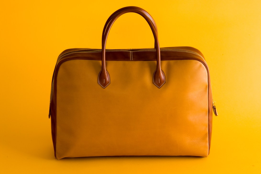 Tan leather handbag with brown handles against a yellow background.