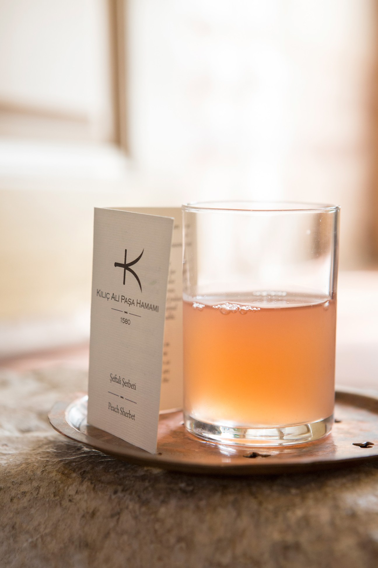 A glass of amber-colored sherbet next to a cream-colored card labeled Kilic Ali Pasa Hamami 1580 on a stone surface.