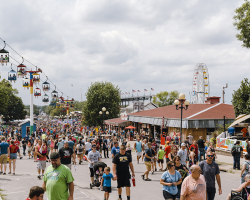 Crowds of fairgoers walking along the main avenue at Iowa State Fair with vendors, rides and attractions visible.