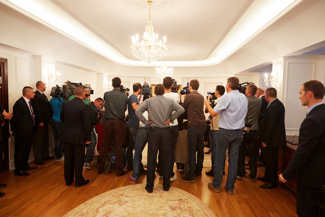 Edi Rama surrounded by press and cameramen in an ornate room with crystal chandeliers during a press meeting in Pristina.