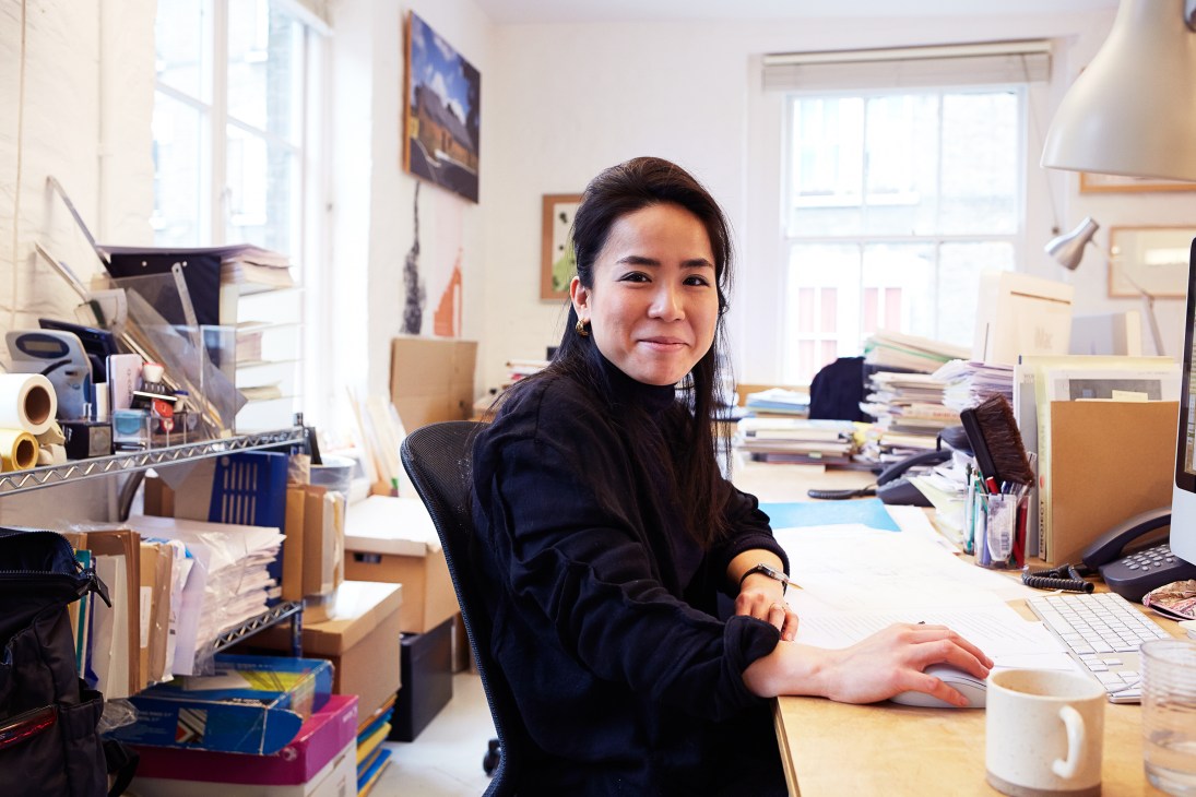 Erika Suzuki sits at her desk in a bright, naturally-lit studio workspace surrounded by books, materials and design references.