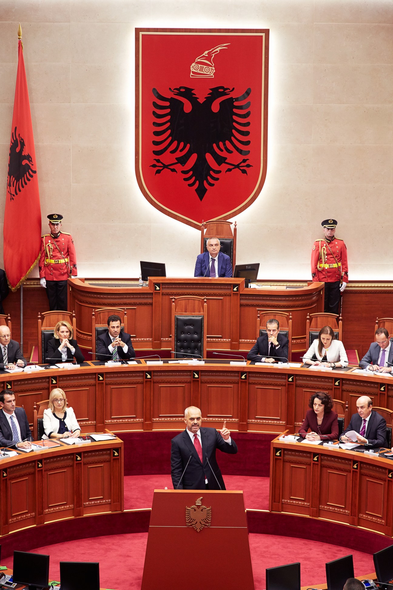 Edi Rama speaks at a podium in Albania's parliament chamber with the Albanian flag and double-headed eagle emblem behind him.