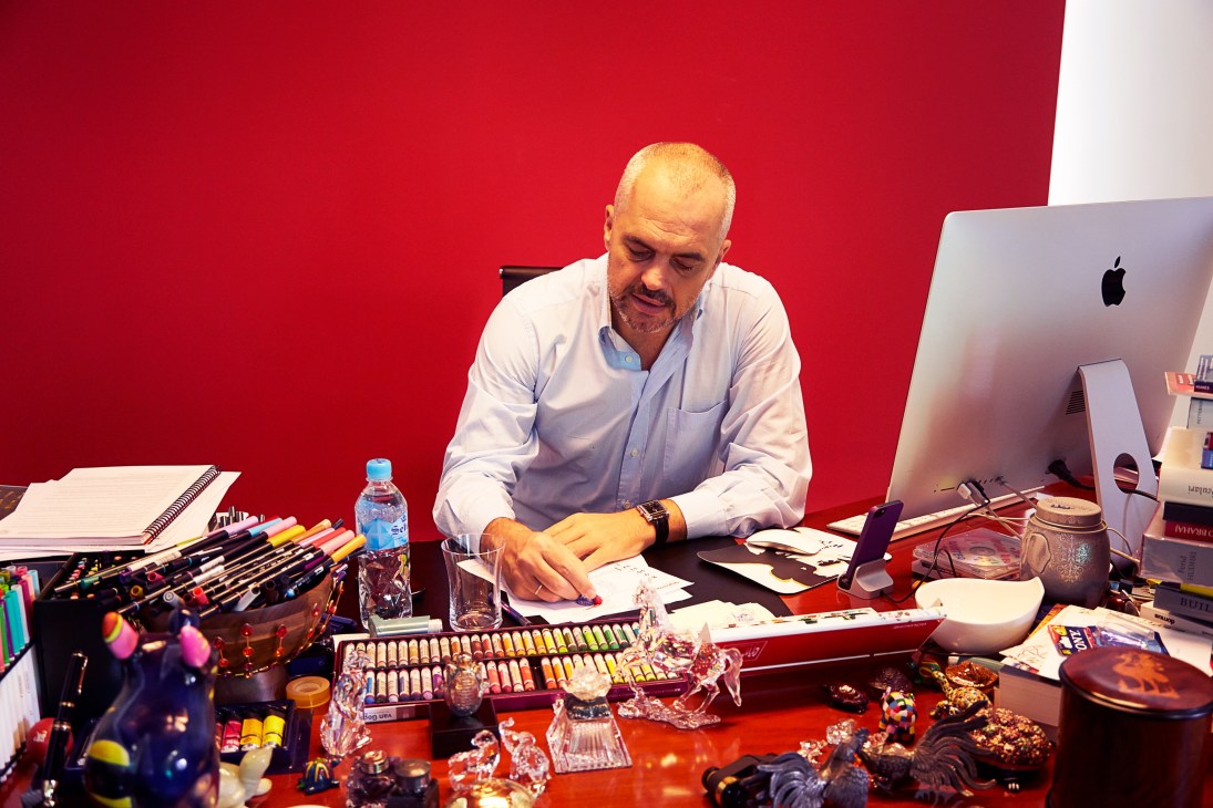 Edi Rama sitting at his desk surrounded by art supplies, sculptures and creative objects against a red wall.