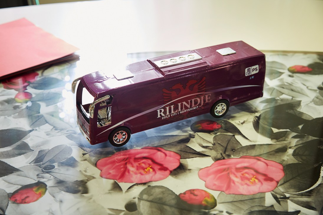 A burgundy model campaign bus labeled 'BLINDJE' sits on a floral patterned surface next to a pink notebook.