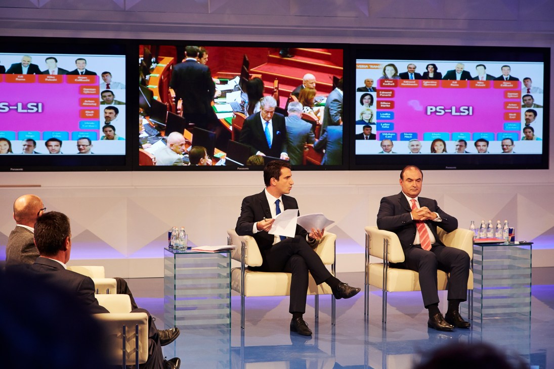 Erion Veliaj sits on a panel at a TV studio with large screens displaying parliament behind him.