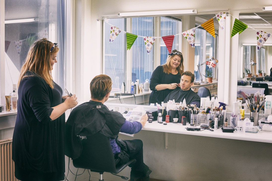 Actor Johannes Lassen sits in a makeup chair while a stylist applies cosmetics in a bright dressing room.
