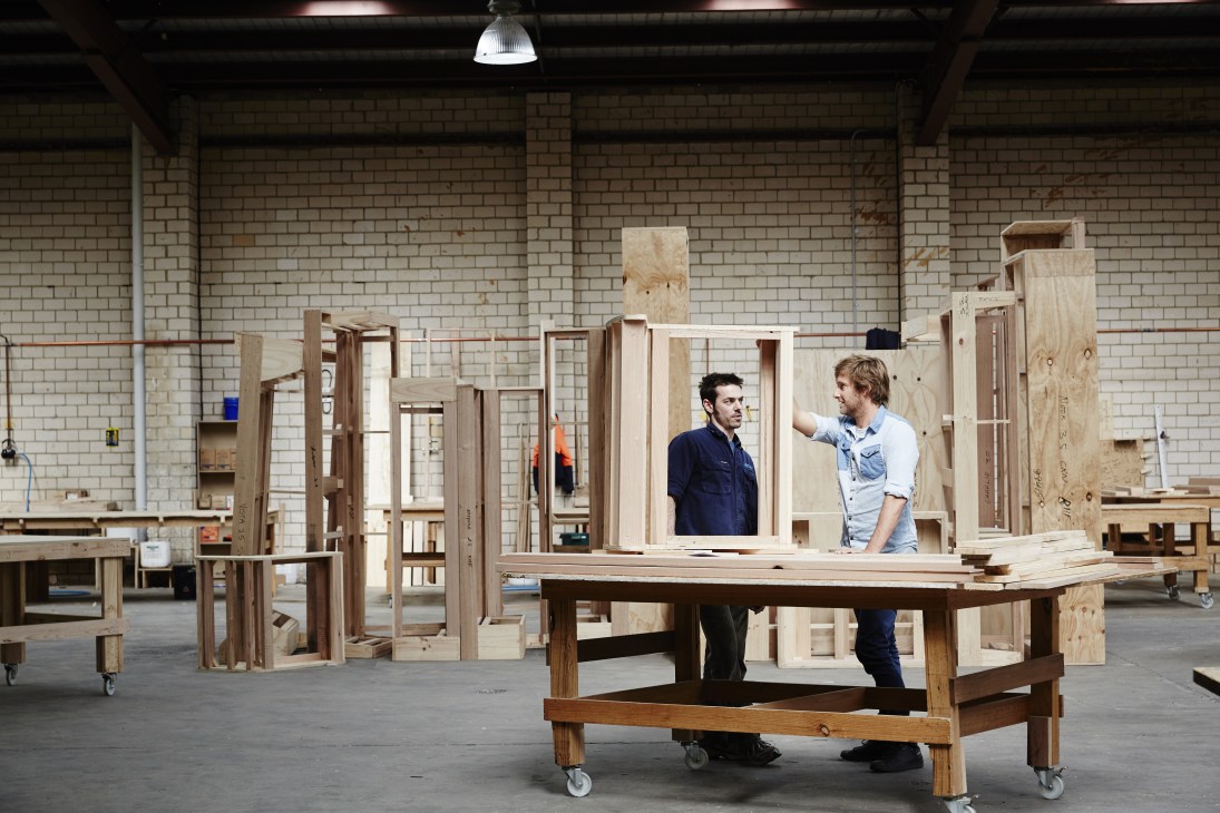 Two workers discuss furniture frames in a Jardan factory workshop filled with wooden chair and table components.