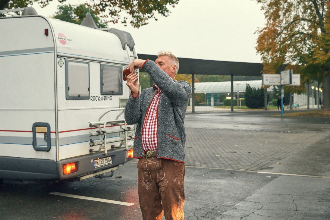 Man in lederhosen and grey jacket adjusting his hat next to a caravan at a campground.