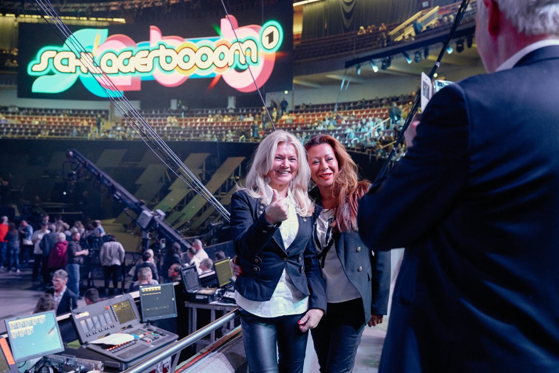 Two women smiling at the Schlagerbooom concert with the neon sign visible above the crowd in Dortmund's Grosse Westfalenhalle.