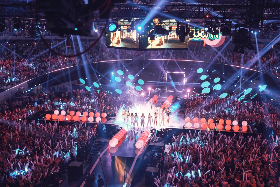 A massive concert stage at Schlagerbooom with performers surrounded by colorful lighting, giant spheres, and thousands of cheering audience members.