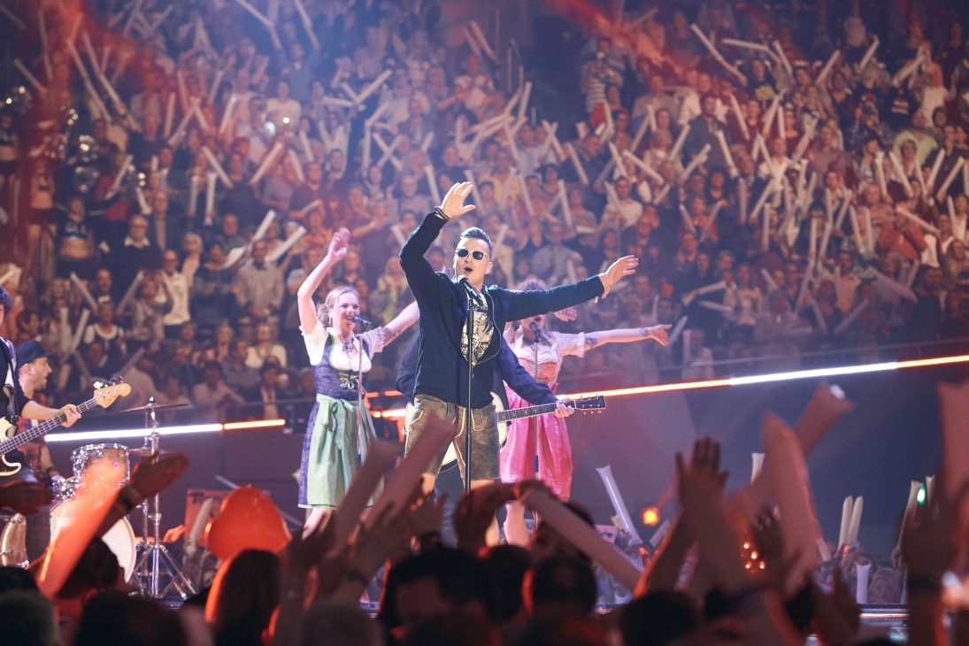 Andreas Gabalier performs on stage wearing sunglasses and lederhosen, arms outstretched to an enthusiastic crowd at Schlagerbooom.