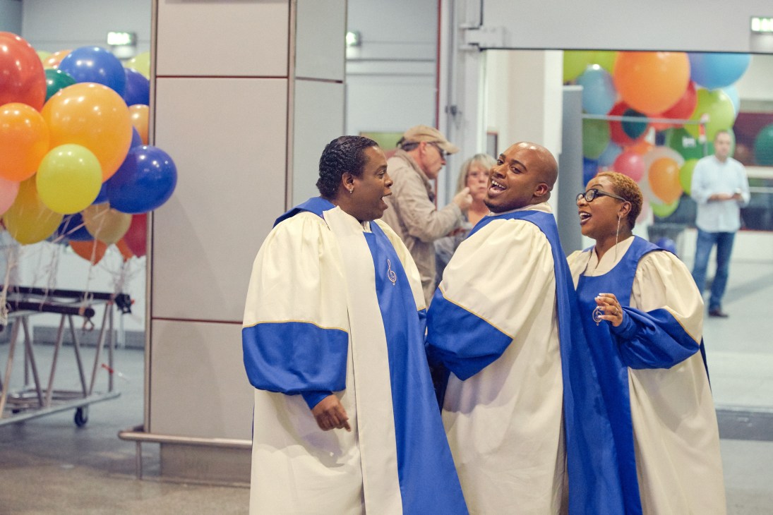 Gospel singers in blue and white robes laughing together at a party with colorful balloons in background.