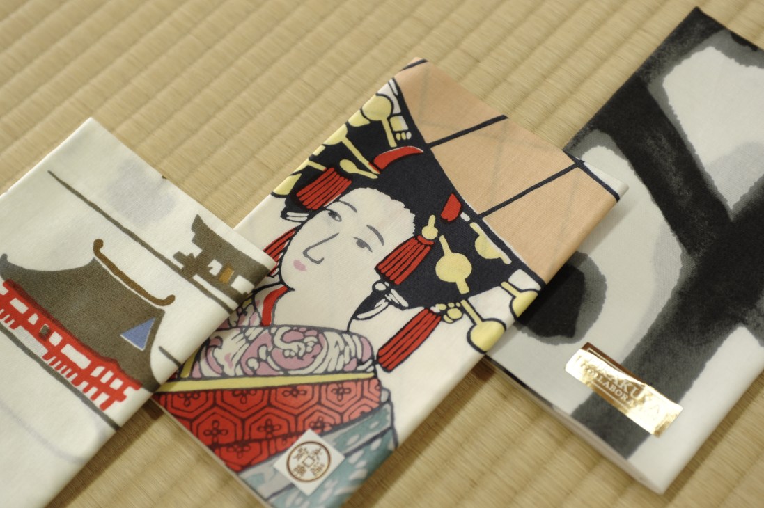 Rolled printed tenugui cloths featuring traditional Japanese designs including a figure in colorful kimono and architectural motifs.