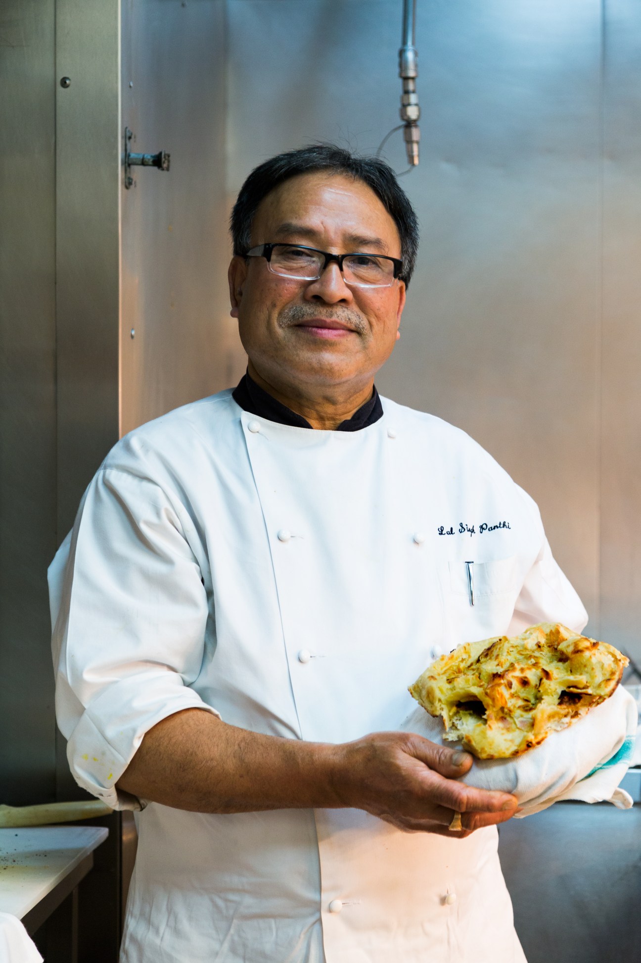 Executive chef Prahlad Hegde in white chef's coat holding a prepared dish in the kitchen.