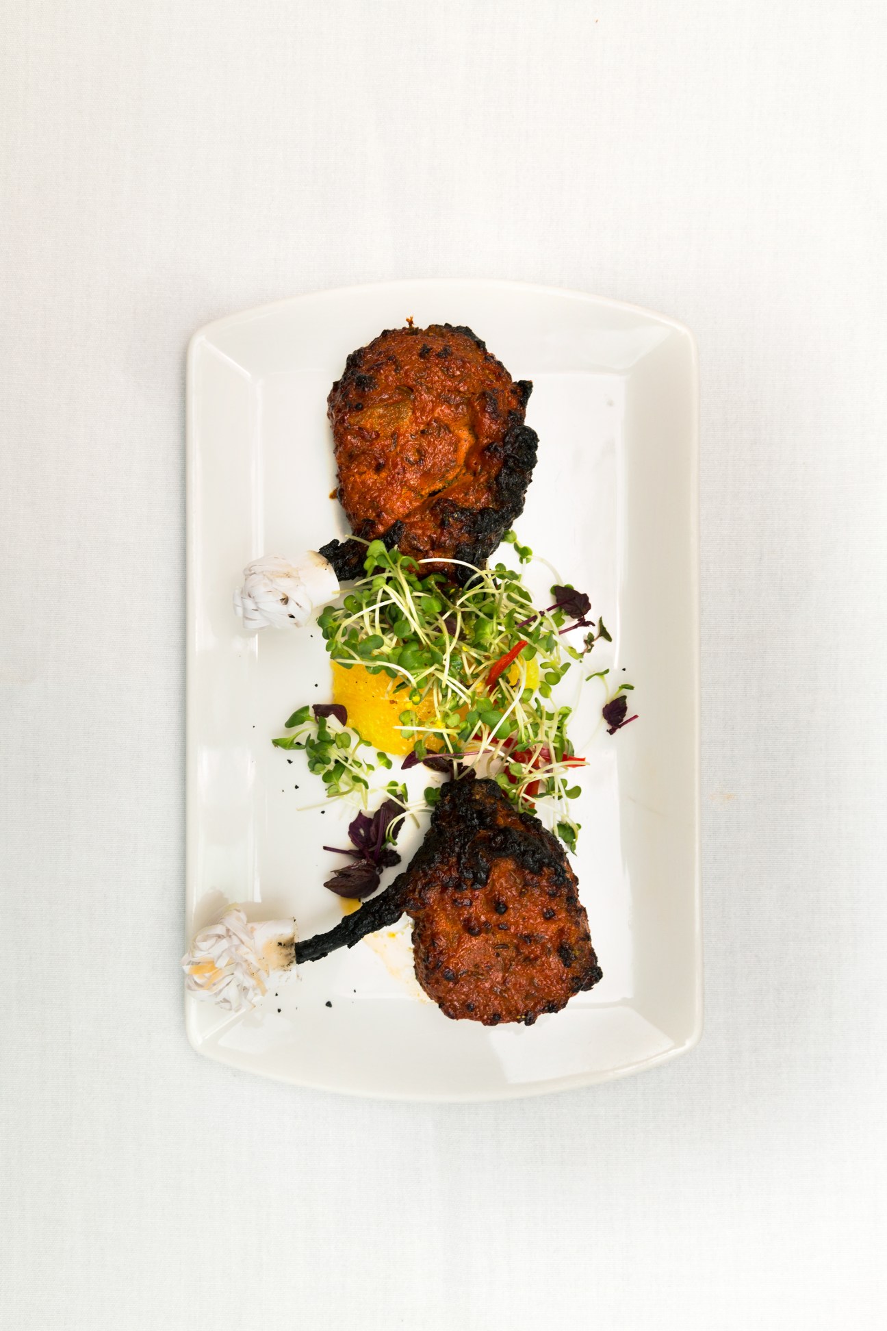 Two tandoori lamb chops with spiced crust served on microgreens and yellow sauce on white plate.