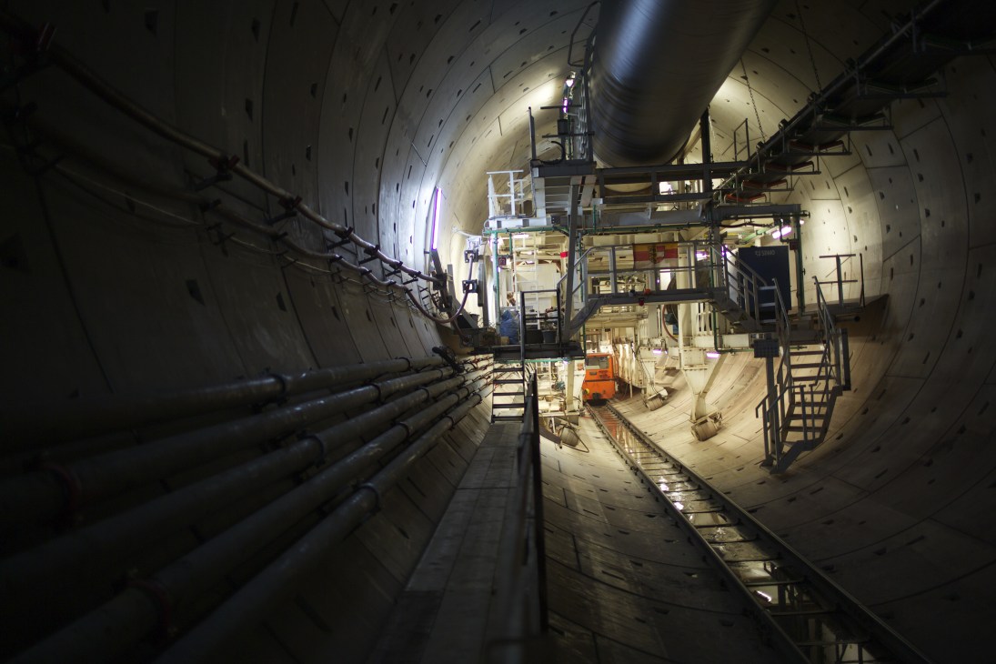 Interior of a large concrete tunnel under construction with pipes, scaffolding and equipment.
