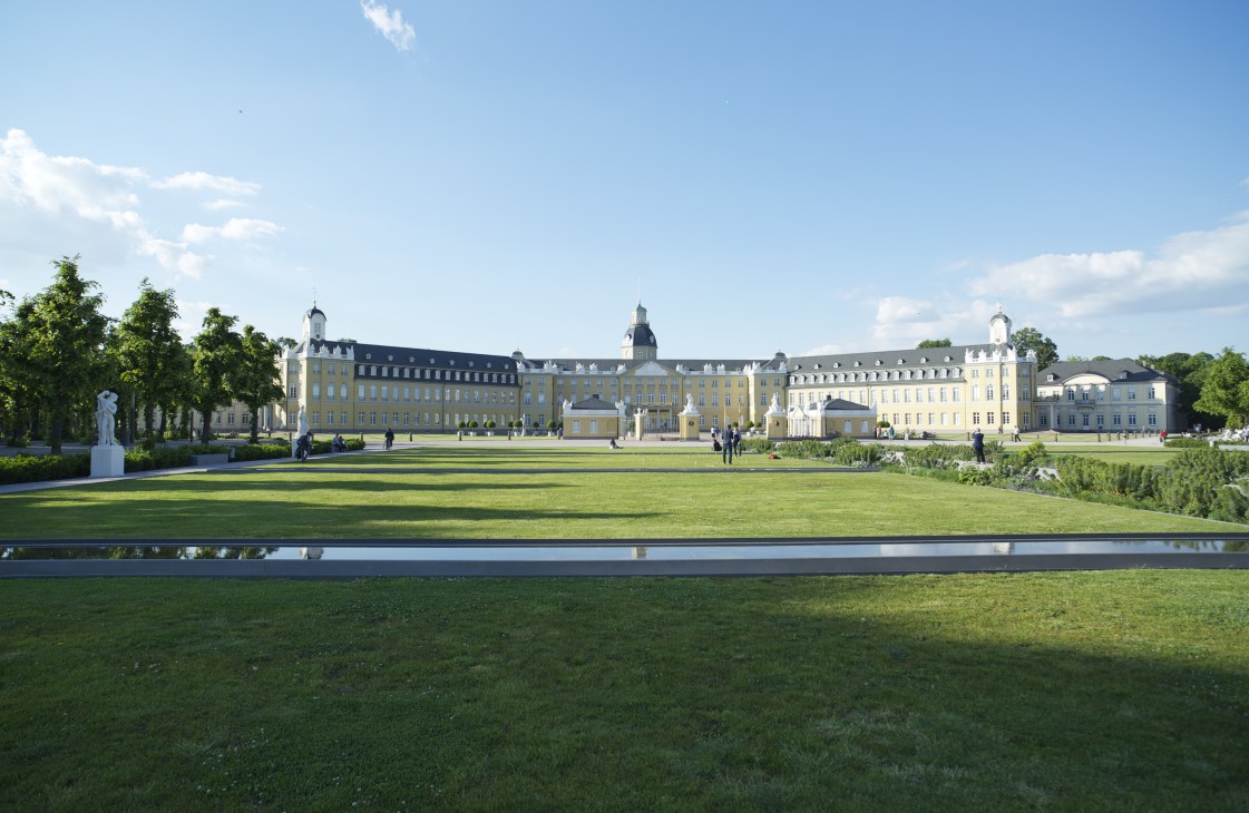 Karlsruhe Palace, a grand yellow neoclassical building with mansard roofs set behind manicured lawns and a water feature.