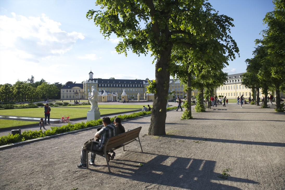 Karlsruhe Palace and tree-lined plaza with visitors and benches on a sunny day.
