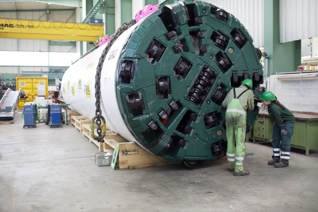 Workers in safety gear inspecting a large green tunnel boring machine component at Herrenknecht factory.