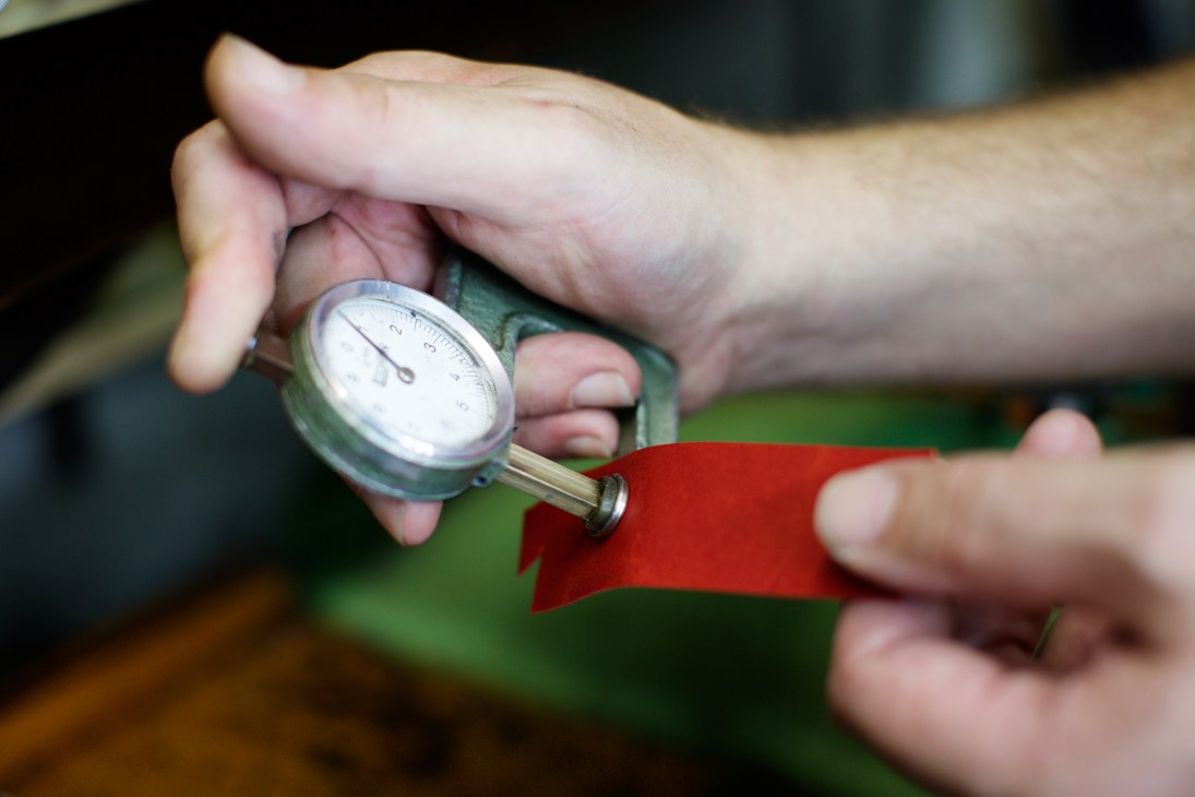 Hands holding a gauge measuring leather thickness, with a red leather sample visible.