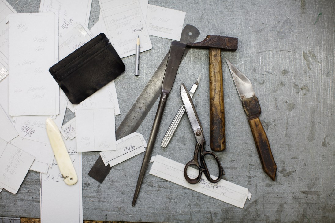 Leather crafting tools and sketches arranged on a gray surface, including a black leather wallet, hammers, knives, scissors, and design drawings.