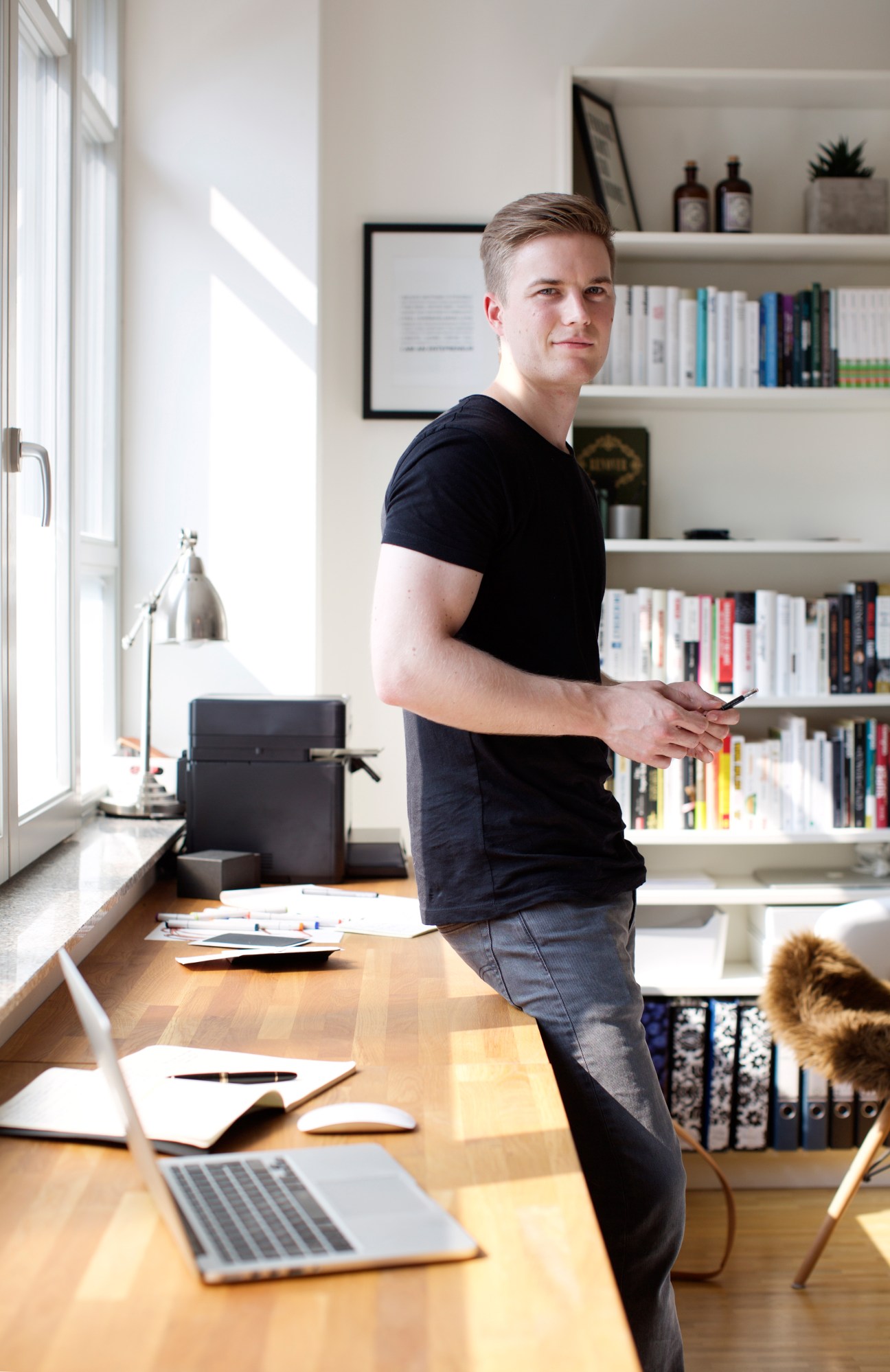 Leander Riedl stands in his Frankfurt office next to a wooden desk with a laptop, wearing a dark navy t-shirt.