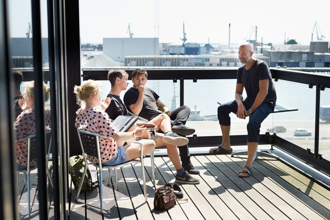 Students and an instructor gather on a rooftop terrace overlooking an industrial port area with cranes and warehouses.