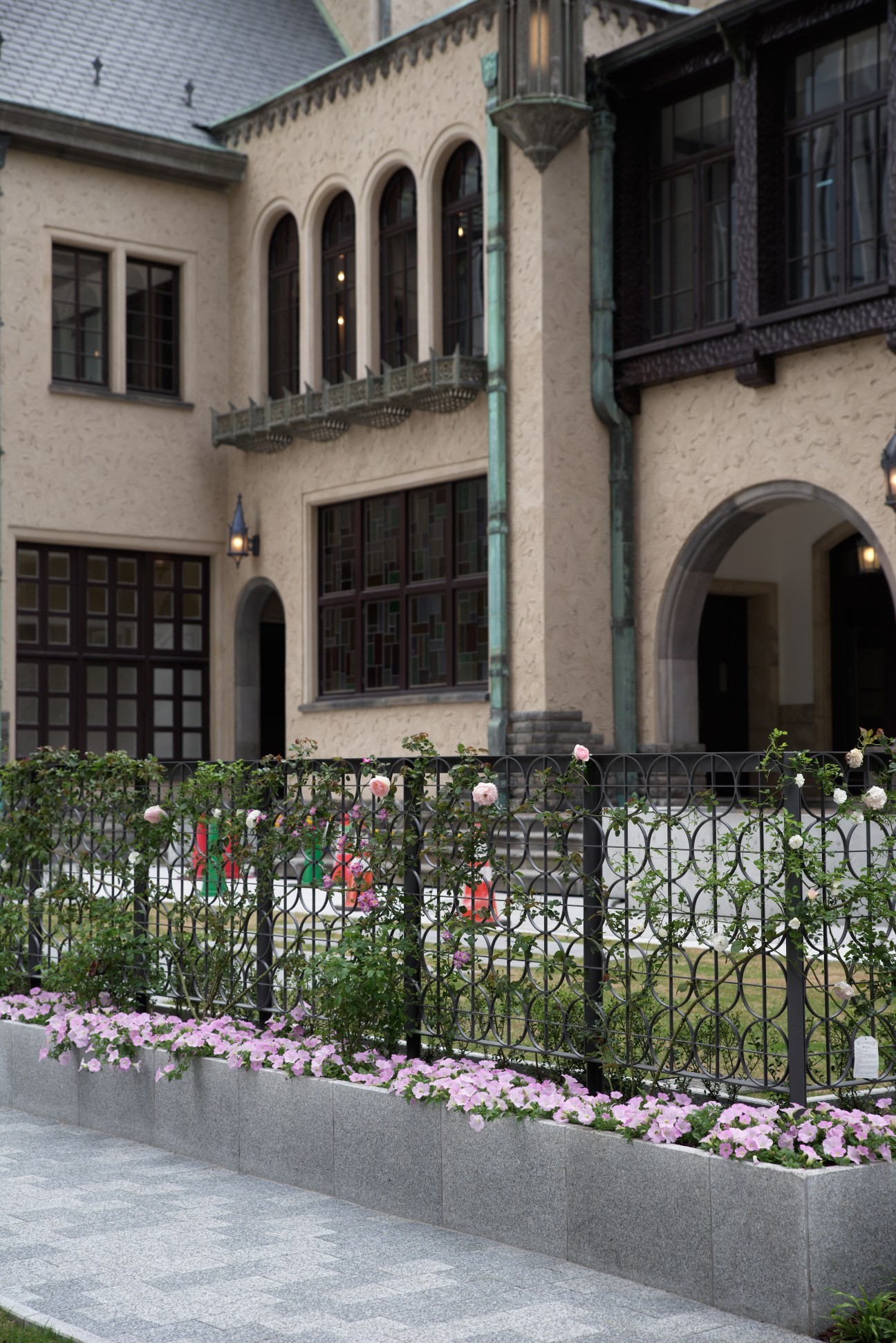 Historic 1930s building facade with arched windows, cream-colored walls, and flowering plants in front garden.
