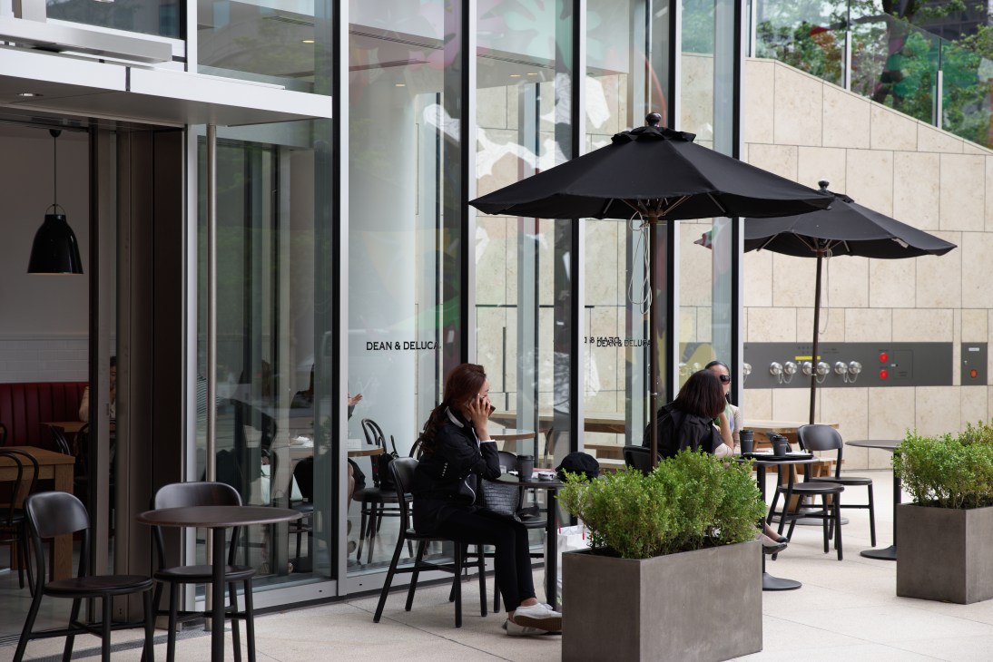 Two women sit at a table with black umbrellas on an outdoor terrace next to a modern building with glass and stone facade.