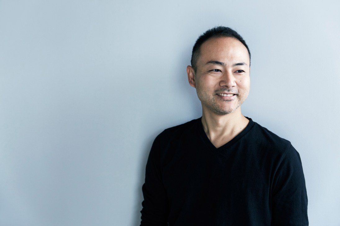Keiji Ashizawa, founder of Ishinomaki Laboratory, smiling at camera against light blue background.