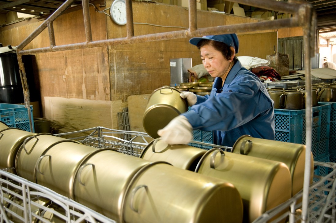 Worker in blue uniform and cap preparing cream-colored steel containers for enamelling at Noda Horo factory.