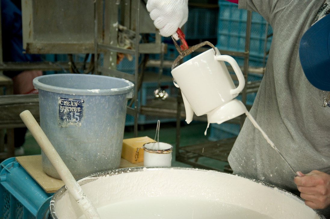 Worker in white gloves coating enamelware pieces multiple times during Noda Horo manufacturing process.