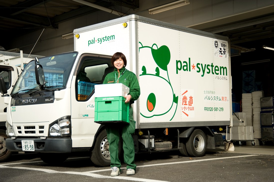 Pal System delivery worker in green uniform holding green crate in front of branded truck with mascot.