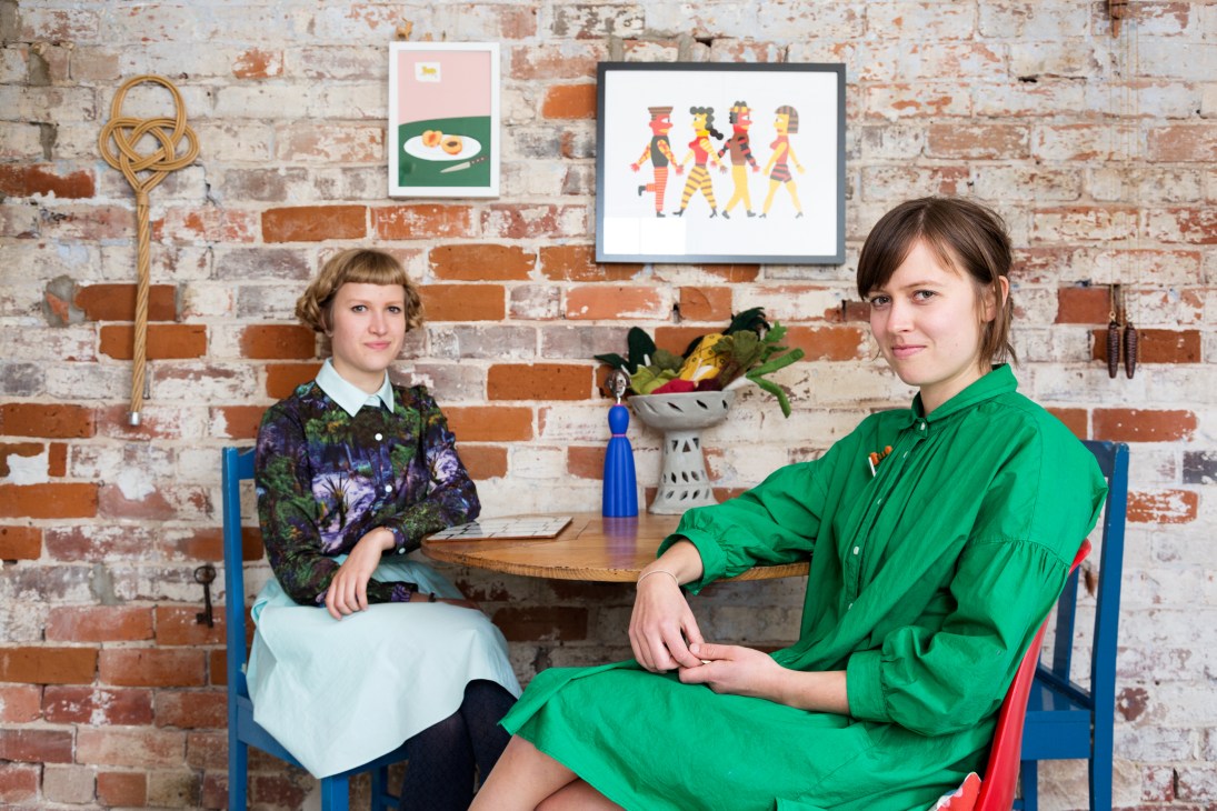 Two women sit at a table against an exposed brick wall decorated with framed artwork and a plant.