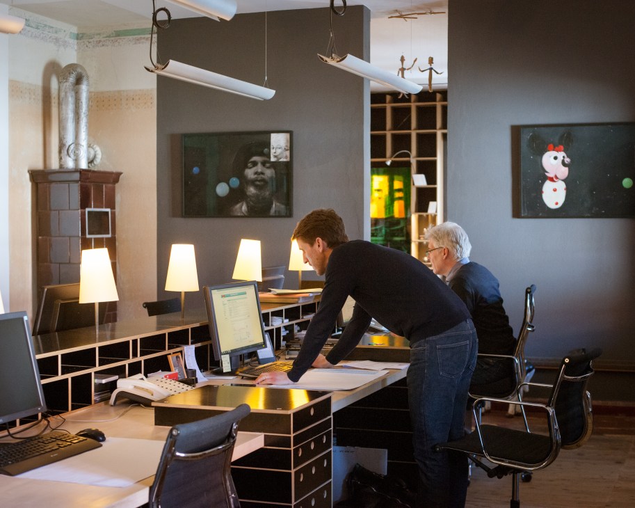 Two people work at a desk in a modern office space with contemporary furniture and hanging lights.