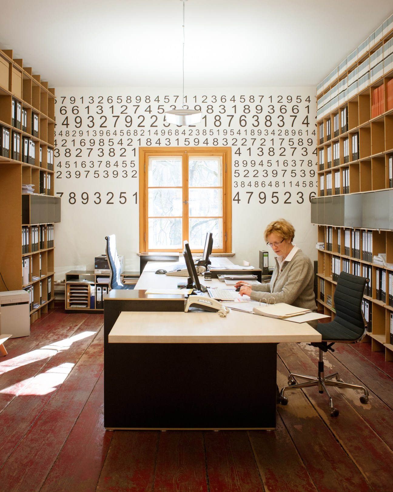 Woman working at desk in office surrounded by wooden bookshelves and a wall decorated with numbers.