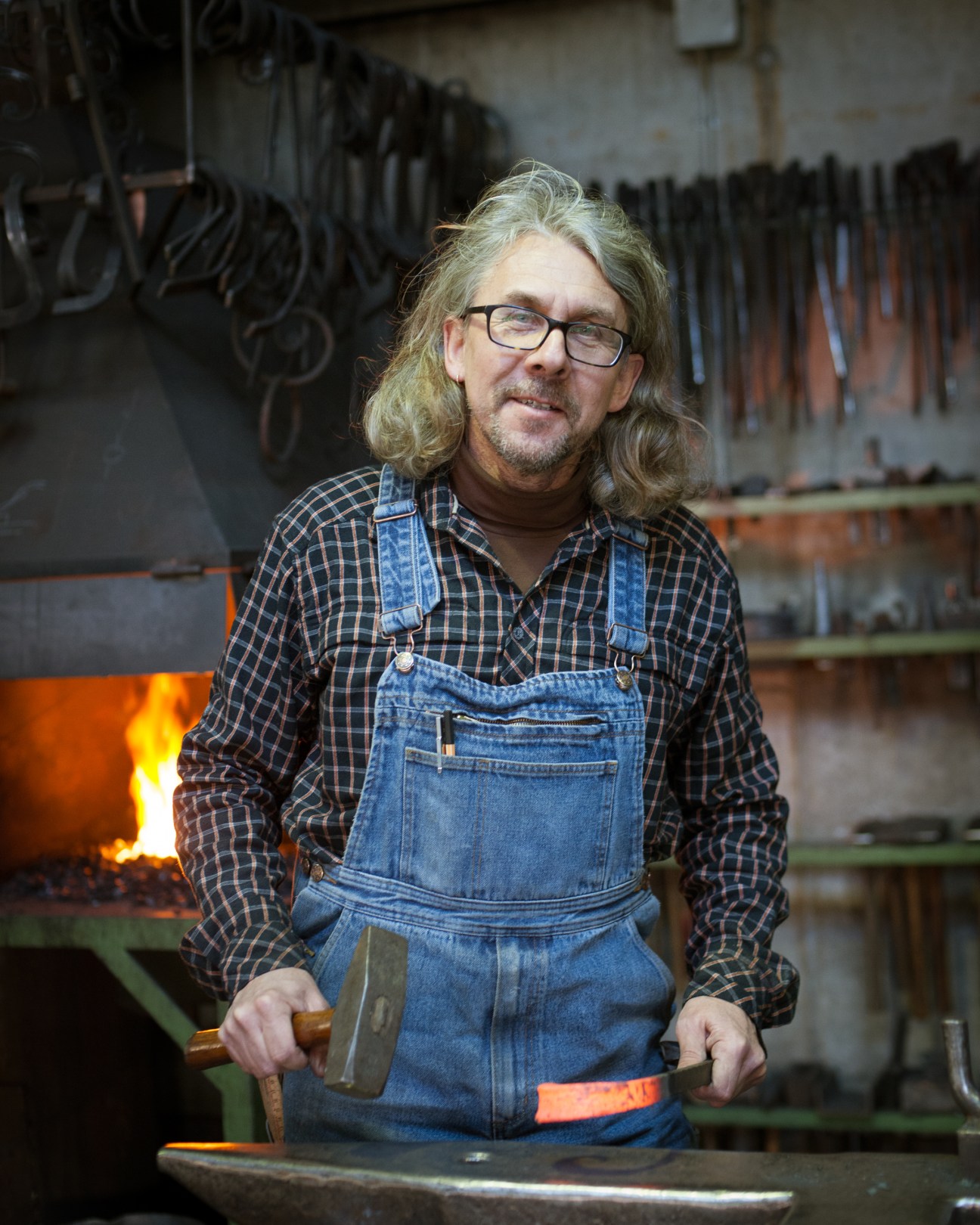 Blacksmith Thedy Metzler stands at his forge wearing a denim apron and checkered shirt, holding metalworking tools with flames visible in the background.