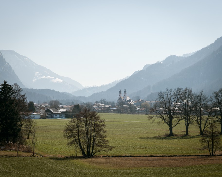 Alpine village of Aschau im Chiemgau nestled in a valley between snow-capped mountains with church spires visible.