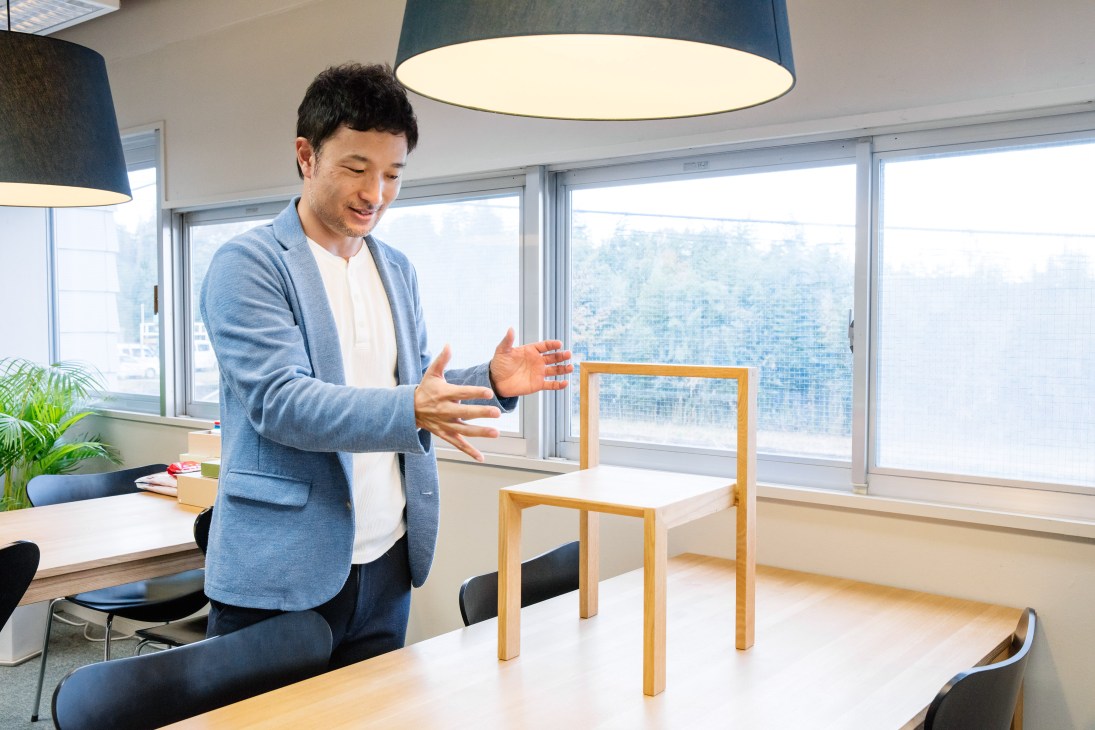 Taku Hibino, architect and designer, gestures while speaking in a bright office with large windows overlooking a cityscape.