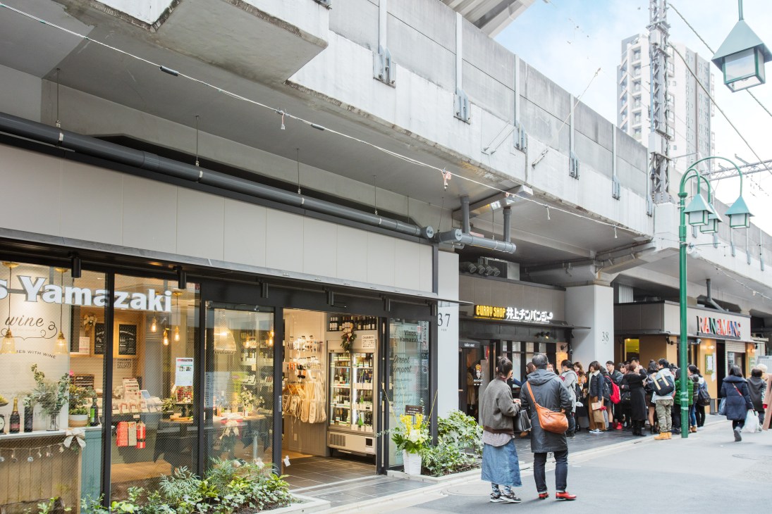 Customers queue outside Inoue Chimpanzee restaurant under Nakameguro Station's railway arches, with other shops visible along the street.