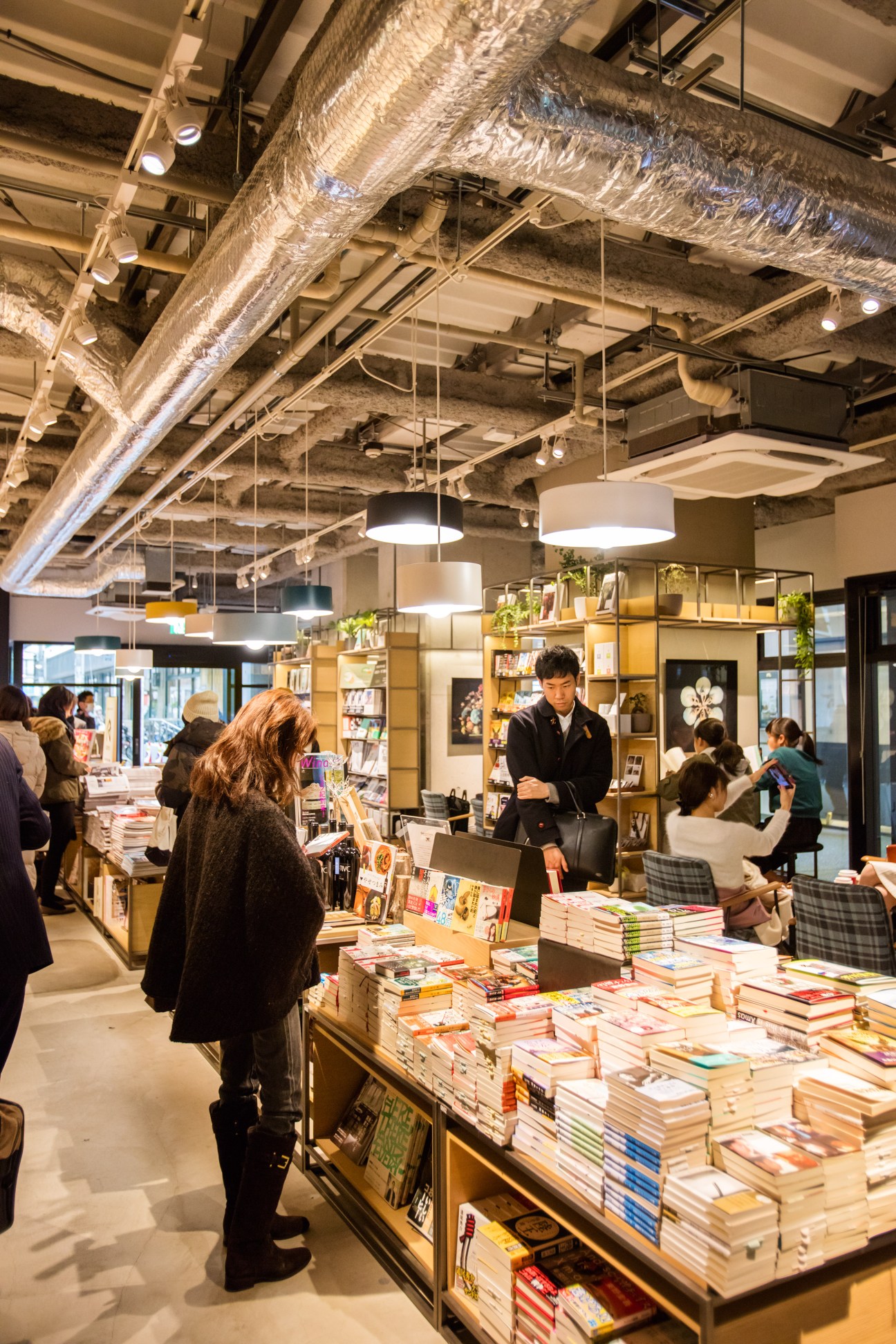 Interior of Tsutaya Books at Nakameguro Station with exposed industrial ceiling, customers browsing books displayed on tables and shelves.