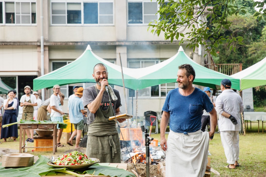 Two chefs, Shin Harakawa and Jerome Waag, speak at an outdoor event under green and white tents with food and attendees visible in the background.