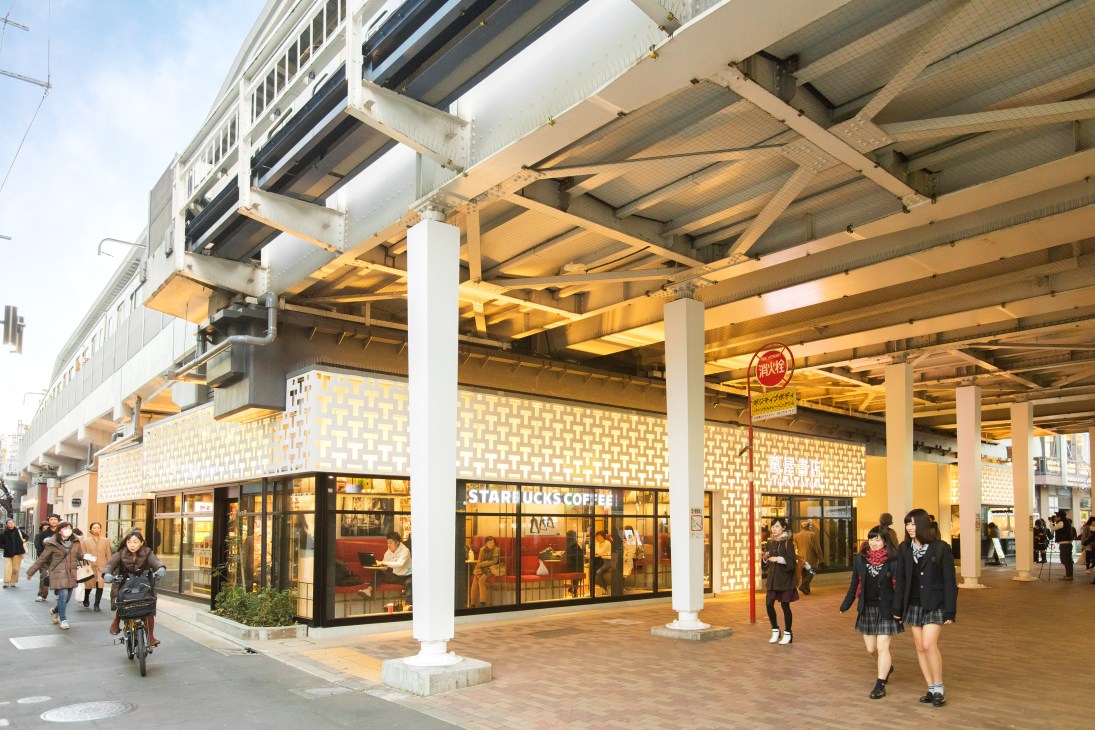Elevated train tracks above a pedestrian shopping area with storefronts and people walking beneath at Nakameguro Station.