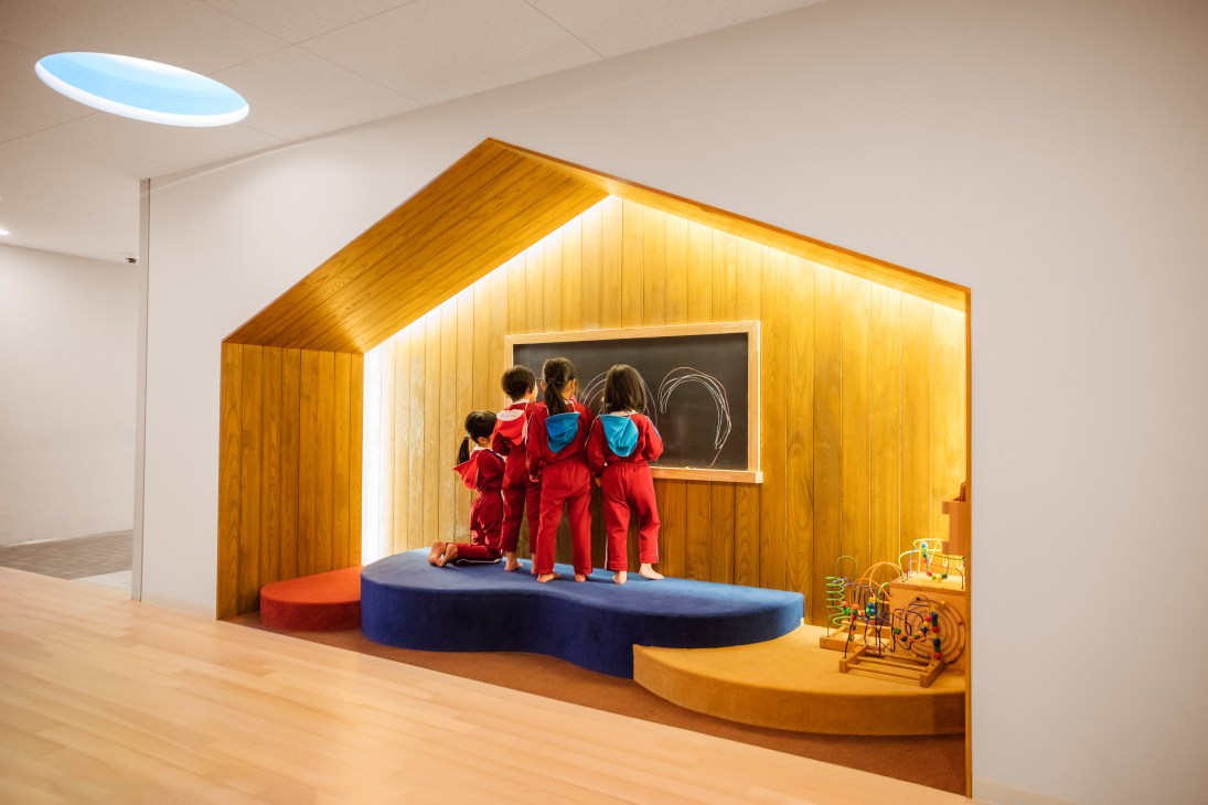 Children in red clothing stand in a wooden alcove with a chalkboard at Obama Kodomoen kindergarten.
