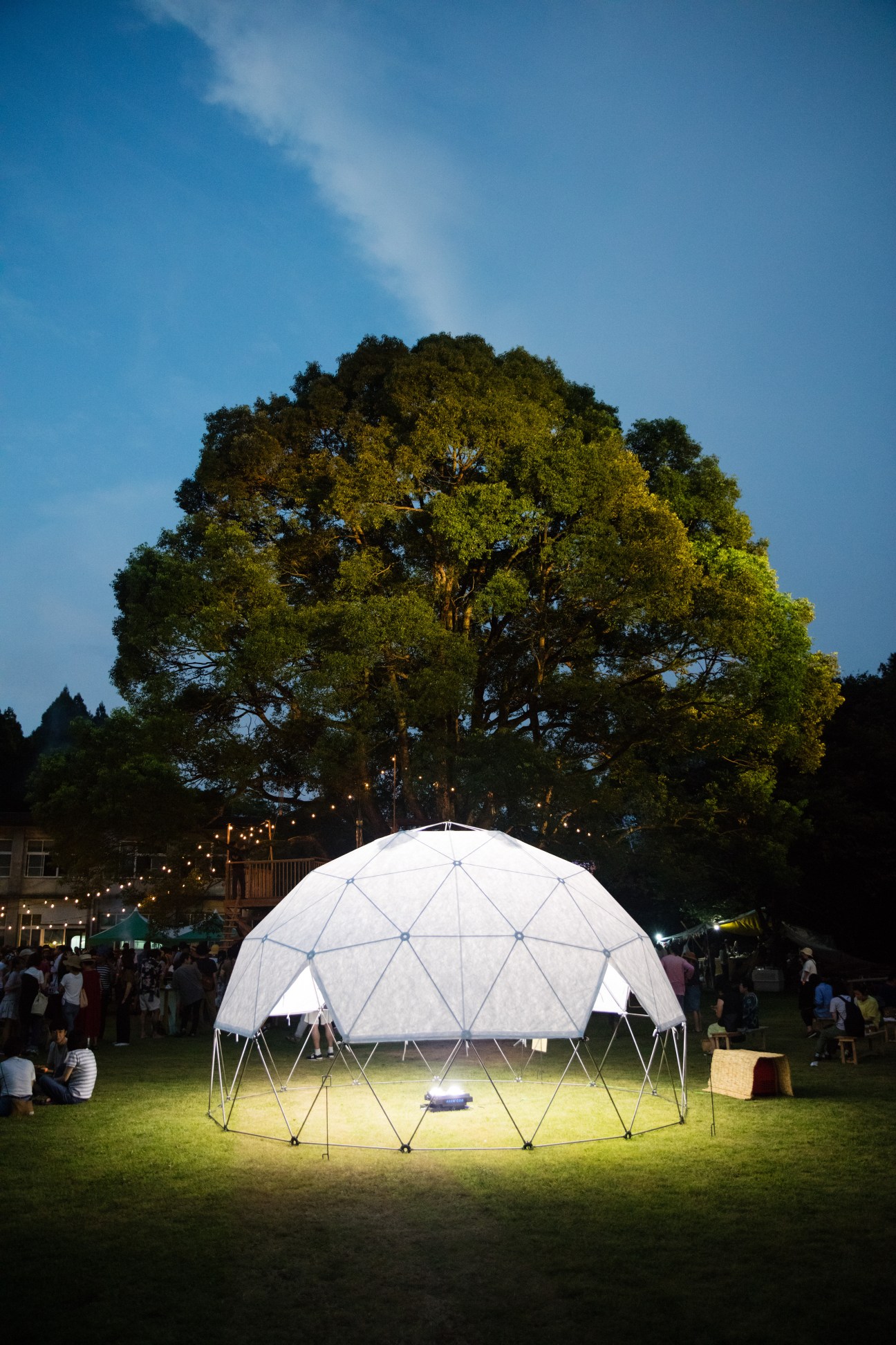 Illuminated white geodesic dome tent at dusk with large tree and crowd beneath string lights at outdoor festival.