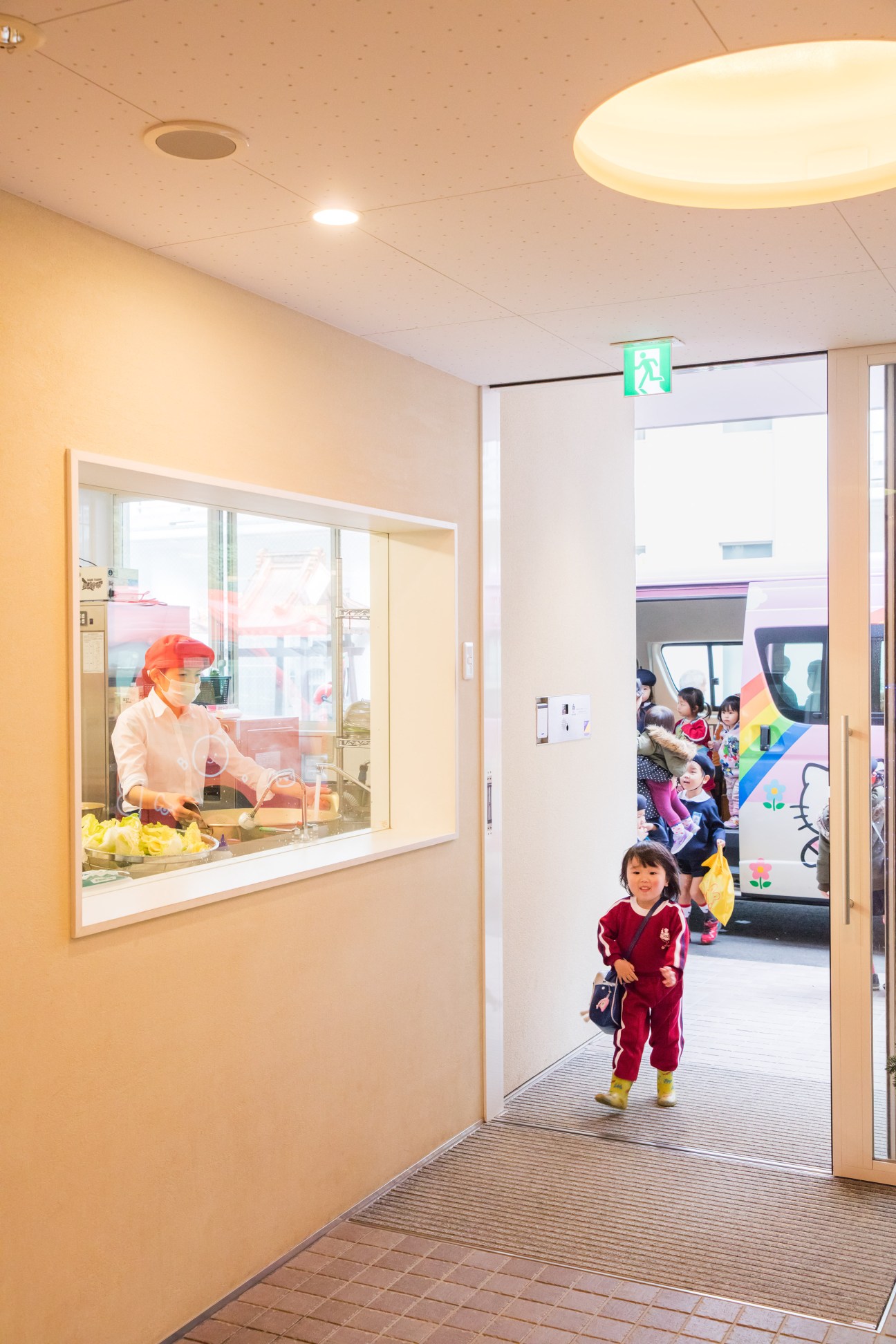 A young child in red clothing runs through a bright corridor with a window showing a chef preparing meals in the kitchen at Obama Kodomoen kindergarten.