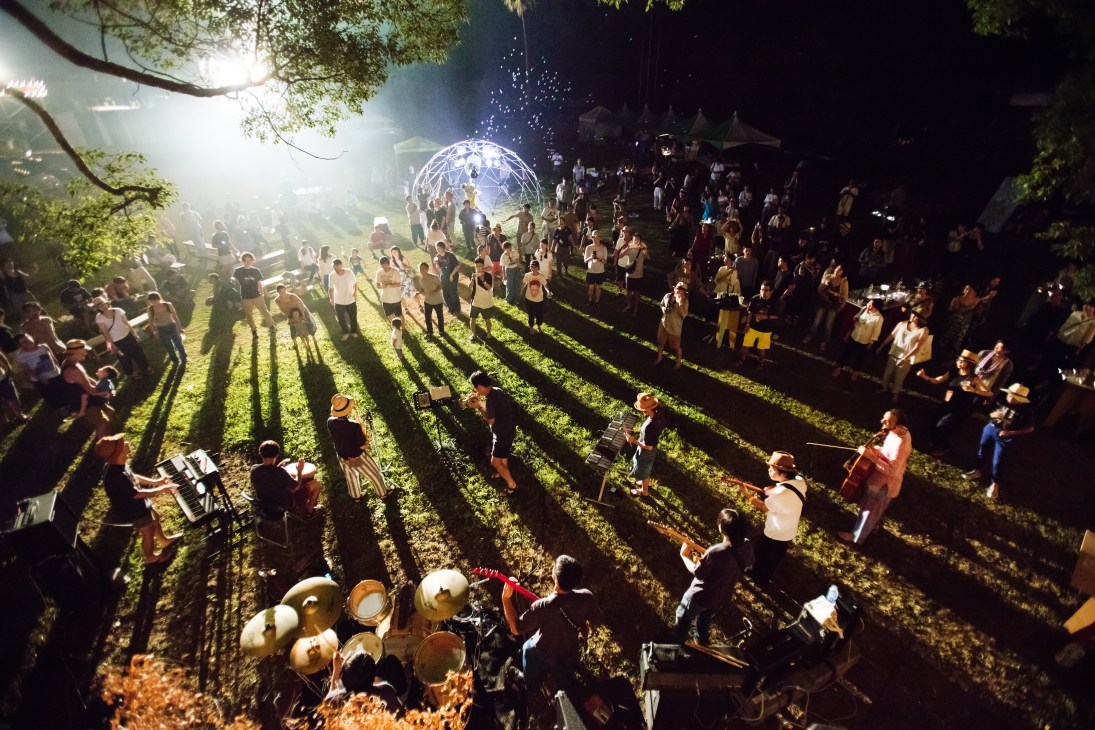 A nighttime outdoor festival scene with crowds gathered around musicians performing on a grass field, illuminated by bright lights and a decorative globe structure.