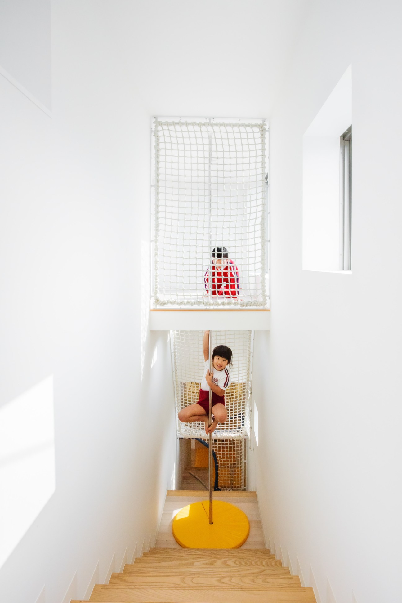 Child climbing on a white net suspended between two levels in a bright stairwell with wooden flooring.
